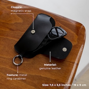 May include: Black leather sunglasses case with a magnetic snap closure and metal ring carabiner. The case is open, revealing a pair of sunglasses. The case measures 7.5 x 3.5 inches.