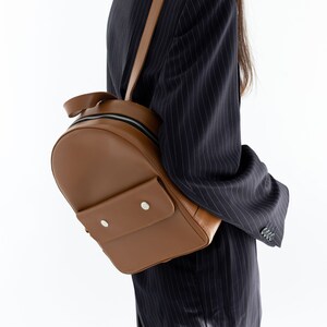 May include: A brown leather backpack with a rounded shape and a small front pocket secured by two silver snaps. The backpack has a top handle and adjustable straps. The zipper is black.