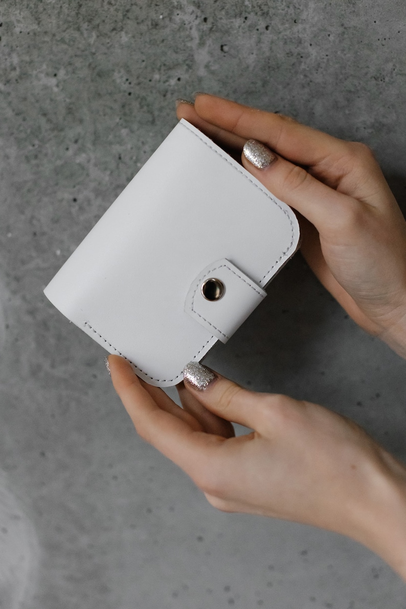 Pu&ograve; includere: Un portafoglio in pelle bianca con chiusura a scatto. Il portafoglio &egrave; tenuto da due mani con smalto glitterato. Il portafoglio ha una forma arrotondata ed &egrave; cucito con un filo a contrasto. Lo sfondo &egrave; grigio screziato.