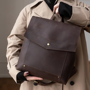 Może przedstawiać: Ciemnobrązowy skórzany plecak z klapą i złotym guzikiem. Plecak ma prostokątny kształt z górnym uchwytem i regulowanymi paskami. Torba jest trzymana przez osobę ubraną w beżowy trencz i czarny sweter.