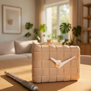 Peut inclure: Une horloge carrée beige tressée avec des aiguilles blanches et un centre doré est posée sur une table en bois. Un stylo argenté se trouve à gauche de l'horloge. L'arrière-plan comprend un canapé, une fenêtre et des plantes.