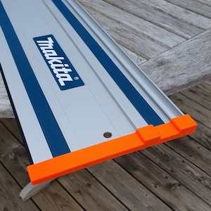 Op de afbeelding: Een zilveren en blauwe meetgids van het merk Makita met een oranje beschermende rand.