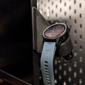 Puede incluir: Un reloj inteligente negro con una correa azul claro está montado en un soporte negro. La esfera del reloj muestra la fecha y la hora. El soporte está fijado a una superficie negra perforada.