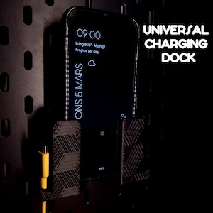 Może przedstawiać: Czarna uniwersalna stacja ładująca z telefonem wyświetlającym godzinę i datę. Stacja ma geometryczny wzór i żółty kabel ładujący. Na zdjęciu widnieje napis "UNIVERSAL CHARGING DOCK".