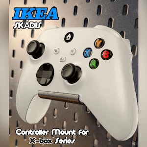 Puede incluir: Soporte blanco para mando de Xbox Series. El soporte está diseñado para sujetar un mando y está montado en una placa perforada. El mando tiene joysticks negros y botones de colores. El texto "IKEA SKÅDIS" y "Controller Mount for X-box Series" son visibles.