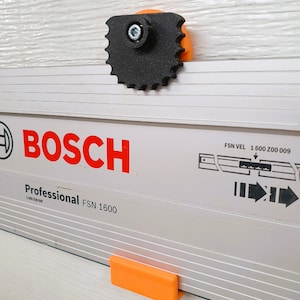 Puede incluir: Un componente negro en forma de engranaje con un perno plateado está fijado a un soporte naranja, montado en un riel guía Bosch Professional FSN 1600 plateado y blanco. El logotipo de Bosch es rojo.