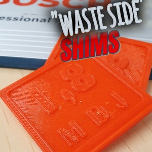 Puede incluir: Dos calzas de plástico naranja con el texto "1/8" y "M.D.J." impreso en ellas. Las calzas están etiquetadas como "Waste Side Shims".