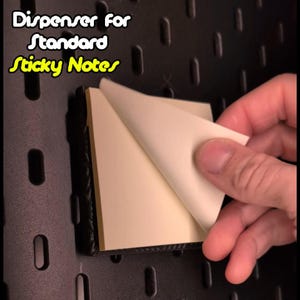 Pode incluir: Um dispensador preto para notas adesivas padrão. O dispensador está montado em uma parede de placa de pinos preta. Uma mão está descolando uma nota adesiva do dispensador.