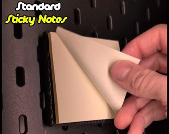 May include: A black dispenser for standard sticky notes. The dispenser is mounted on a black pegboard wall. A hand is peeling a sticky note from the dispenser.
