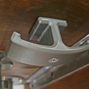 Puede incluir: Un gancho gris impreso en 3D con una base en forma de T. El gancho está fijado a una superficie de madera con un tornillo.