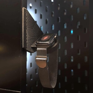 May include: A black smartwatch with a silver strap is displayed on a black 3D-printed holder. The watch face shows a red lightning bolt symbol. The holder is mounted on a dark, perforated wall.