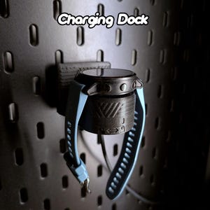 Könnte beinhalten: Eine schwarze Ladestation mit einem blauen Uhrenarmband. Die Ladestation hat eine strukturierte Oberfläche und der Text "Charging Dock" ist über der Ladestation sichtbar.