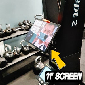 May include: An 11-inch screen is mounted on a black exercise machine. The screen displays a review of a phone. Dumbbells and kettlebells are visible on shelves in the background.