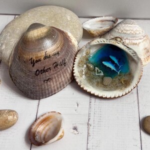May include: Two decorated seashells. One shell has the text "You are my Other Half" and an infinity symbol. The other shell has a blue resin scene with two dolphins, sand, and a heart symbol. Other seashells and a stone are in the background.