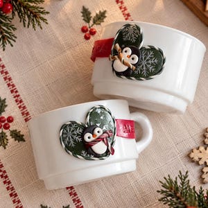 May include: Two white ceramic mugs featuring a decorative heart-shaped emblem on the side. The emblem is dark green with a white snowflake pattern and a small penguin figure wearing a striped scarf. A red band with the text Merry is attached.