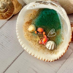 May include: A decorative seashell with a miniature ocean scene. The shell is white with a textured edge, containing a teal and green resin ocean, tiny shells, and small orange crabs. Gold glitter adds sparkle.