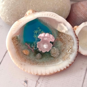 Peut inclure: Une coquille Saint-Jacques décorative avec une scène sous-marine miniature. La coquille contient de la résine bleue, du sable et de petits coquillages. Une coquille de palourde rose avec une perle et du corail sont au centre. La coquille est blanche et beige.