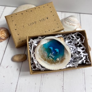 May include: A seashell art piece with a blue ocean scene and two dolphins, presented in a small brown gift box. The box is open, revealing the shell filled with resin, sand, and the words "I love you". The box is decorated with small heart shapes.