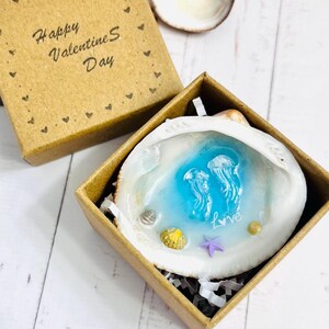 Puede incluir: Una caja de regalo cuadrada de cartón marrón con el texto Happy Valentine's Day impreso en la tapa. Dentro, una concha blanca contiene una piscina de resina azul con dos figuras de medusas, una pequeña estrella de mar morada y conchas.