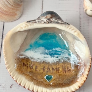 Puede incluir: Una concha marina decorativa con una escena oceánica de resina que muestra olas blancas rompiendo en una playa de arena. La zona de arena incluye el texto grabado MIKE + AMELIA y un pequeño corazón de color verde azulado. Interior color crema.