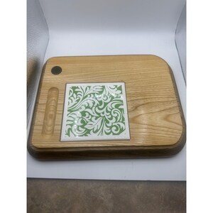 May include: A rectangular wooden serving board with rounded corners, featuring a light brown wood grain finish. A square white ceramic tile with a green leafy scroll pattern is inlaid in the center. A small circular dark inlay and a vertical groove are present.