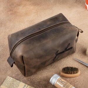 May include: A brown leather toiletry bag with a zipper closure and the name "Jonathan" embossed on the front. The bag is sitting on a brown surface with a brush, scissors, and a tube of hair product.