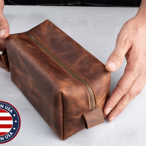 Peut inclure: Une trousse de toilette en cuir marron avec une fermeture &eacute;clair et une poign&eacute;e en cuir. Le sac est fabriqu&eacute; aux &Eacute;tats-Unis.