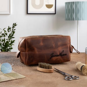 Peut inclure: Une trousse de toilette en cuir marron avec le nom "Jonathan" grav&eacute; sur le devant. La trousse est pos&eacute;e sur une surface en bois avec une brosse, des ciseaux et un r&eacute;cipient de cire.