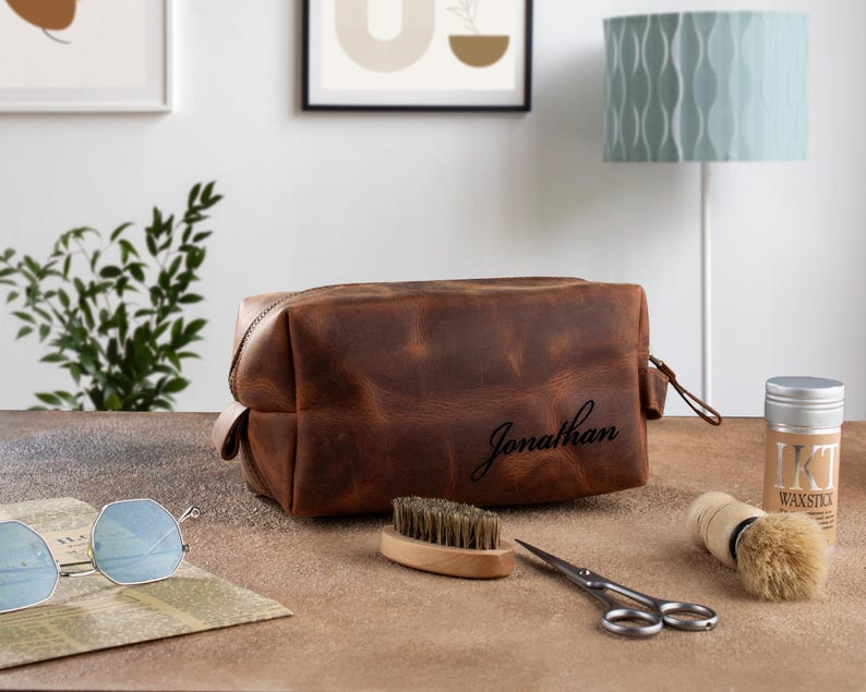 Peut inclure: Une trousse de toilette en cuir marron avec le nom "Jonathan" grav&eacute; sur le devant. La trousse est pos&eacute;e sur une surface en bois avec une brosse, des ciseaux et un r&eacute;cipient de cire.