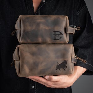 Peut inclure: Deux trousses de toilette en cuir marron avec des fermetures &agrave; glissi&egrave;re. La trousse du haut porte le monogramme "DAVID" sur le devant. La trousse du bas pr&eacute;sente un motif de silhouette de taureau.