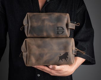 Peut inclure: Deux trousses de toilette en cuir marron avec des fermetures à glissière. La trousse du haut est monogramme avec le nom "DAVID" sur le devant. La trousse du bas présente un motif de silhouette de taureau.