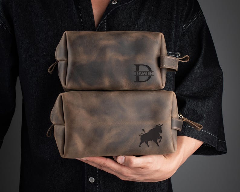 Peut inclure: Deux trousses de toilette en cuir marron avec des fermetures &agrave; glissi&egrave;re. La trousse du haut porte le monogramme "DAVID" sur le devant. La trousse du bas pr&eacute;sente un motif de silhouette de taureau.