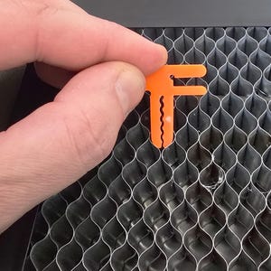 May include: An orange plastic tool with a T-shape and serrated edges is being used to secure a metal honeycomb panel.