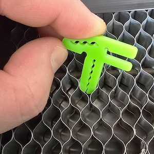 May include: A bright green plastic T-shaped corner brace with a textured surface. The brace is being held over a metal honeycomb panel.