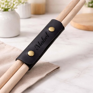 May include: Black leather drumstick holder with the name "Mabel" engraved. The holder has two brass-colored snap closures and holds two wooden drumsticks. The drumsticks are light brown. The holder is resting on a beige cloth.