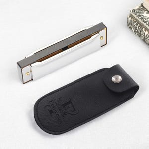 May include: A silver harmonica with a black top and gold accents, next to a black leather case with the name "Francesca" printed on it. The harmonica is a musical instrument, likely for blues or folk music.