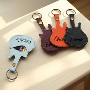 May include: A collection of guitar-shaped keychains in various colors, including purple, orange, and black. Each keychain has a name printed on it. A light blue guitar pick holder with the name "Camilla" is also present.