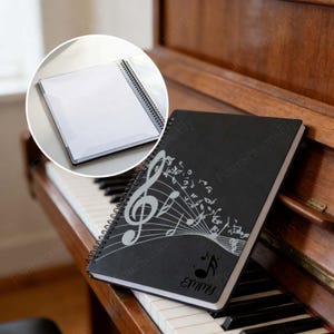 May include: A black spiral-bound notebook featuring a white musical note design. The notebook is personalised with the name "Emmy" and is placed on a piano keyboard. A clear plastic notebook is in the background.