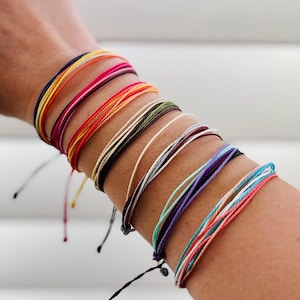 May include: A collection of multiple thin, waterproof string bracelets worn together. The bracelets feature a vibrant array of colors including navy, yellow, orange, pink, red, olive green, black, white, burgundy, purple, teal, and light blue.