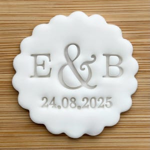 May include: A white, scalloped-edge emblem featuring the letters "E & B" and the date "24.08.2025" in a simple font. The emblem is set against a light brown, wood-grain background.
