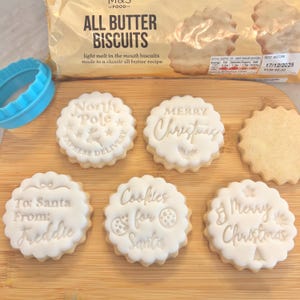 May include: A selection of all butter biscuits with white icing and Christmas-themed messages. The biscuits are round with a scalloped edge and feature text like "Merry Christmas" and "Cookies for Santa". A blue cookie cutter is also visible.