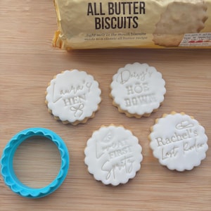 May include: Four iced biscuits with phrases like "Laura's Hen" and "Rachel's Last Rodeo" sit on a wooden surface. A blue, scalloped cookie cutter and a packet of "All Butter Biscuits" are also present.