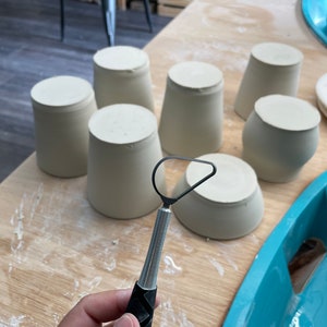 May include: A close-up of a pottery tool with a metal loop and a black handle. The tool is being held by a hand. There are several white ceramic cups on a wooden table.