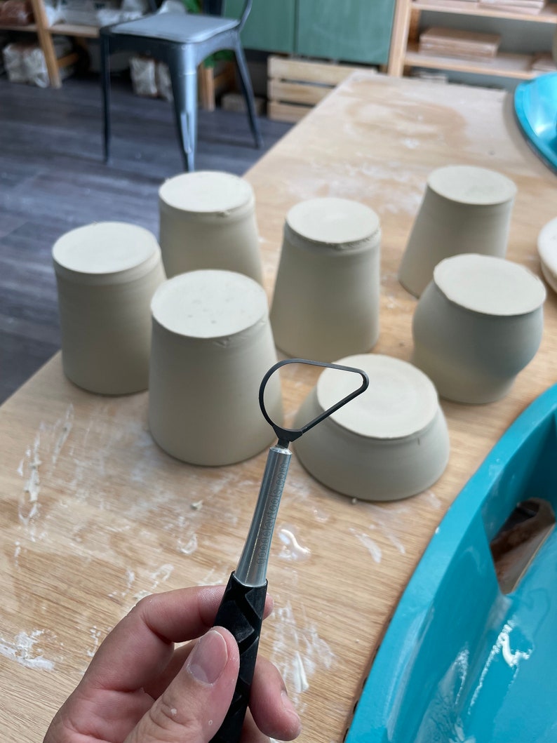 May include: A close-up of a pottery tool with a metal loop and a black handle. The tool is being held by a hand. There are several white ceramic cups on a wooden table.