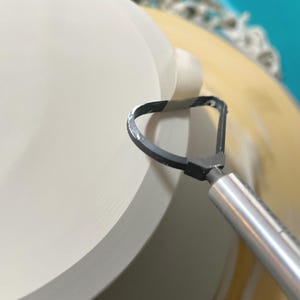 May include: A close-up of a pottery wheel with a ceramic bowl being shaped. A metal tool with a loop is being used to smooth the edge of the bowl.