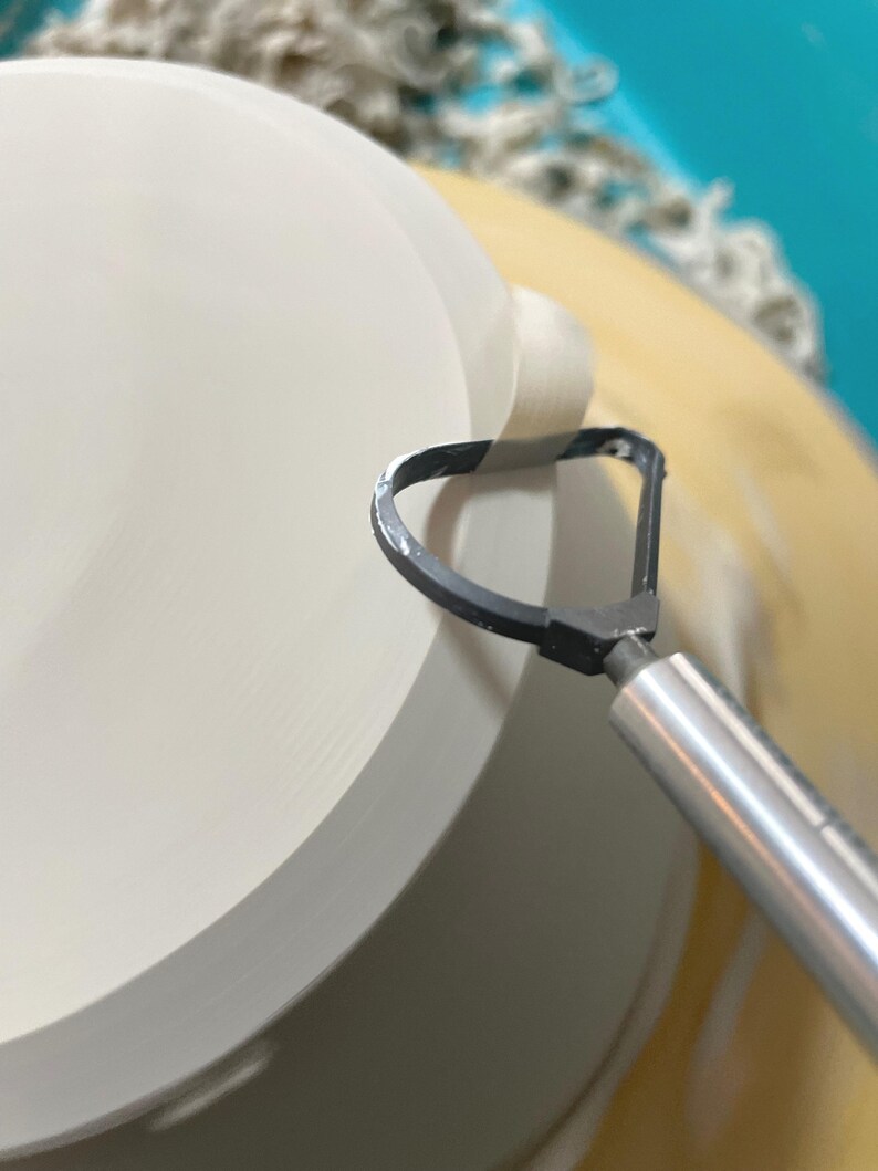 May include: A close-up of a pottery wheel with a ceramic bowl being shaped. A metal tool with a loop is being used to smooth the edge of the bowl.