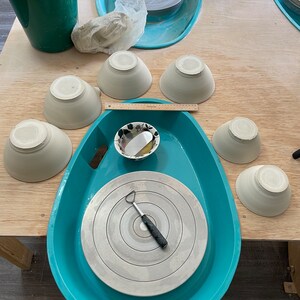 May include: A turquoise pottery wheel with a white ceramic disc and a small metal tool. There are several white ceramic bowls on a wooden table.