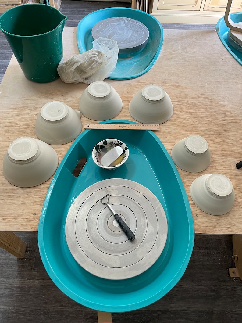 May include: A turquoise pottery wheel with a white ceramic disc and a small metal tool. There are several white ceramic bowls on a wooden table.