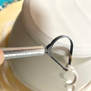May include: A close-up of a silver tool with a black loop on the end. The tool is being used to trim the edge of a white ceramic pot. The tool has the text "claytools.com" engraved on the side.