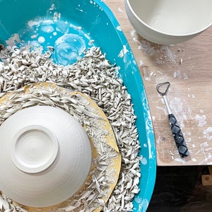 May include: A white ceramic bowl sits on a pottery wheel, surrounded by white clay shavings. A turquoise plastic tray holds the wheel and shavings. A second white ceramic bowl sits on a wooden table next to the tray.